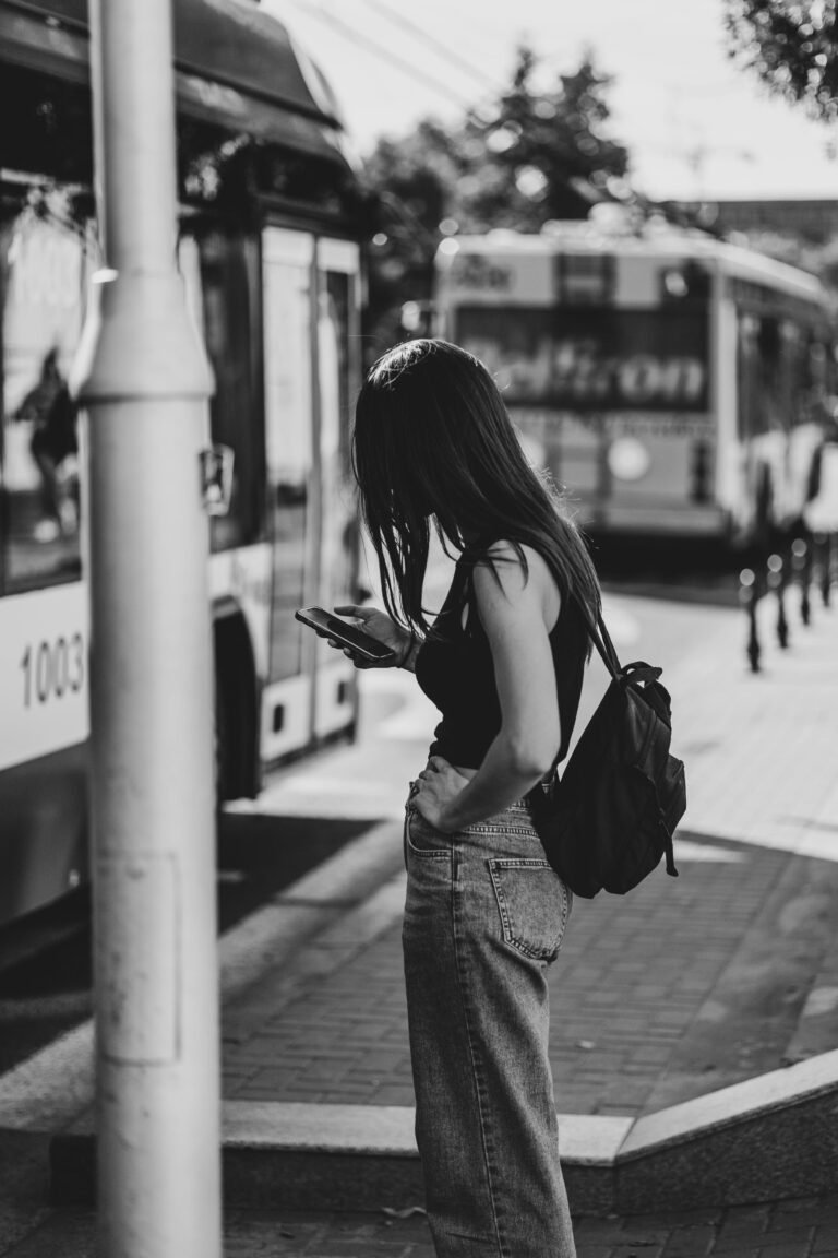 Black and white photo of a woman at a city bus stop, using her phone and wearing casual attire.